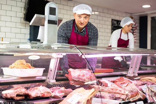 Positive man, professional butcher shop seller, arranging meat products in display case, showing piece of fresh raw veal tenderloin