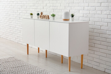 Stylish white chest of drawers with houseplants and decor near light brick wall in room