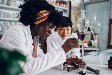 A happy african american pharmacist is using a funnel for cure making while her Japanese colleague...