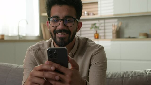 Indian Arabian millennial guy happy smiling man freelancer on couch rest weekend with mobile phone scrolling internet media use smartphone app shopping make online order delivery food at home kitchen