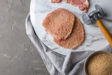 Cooking schnitzel. Raw pork chops in bread crumbs and meat mallet on grey table, top view with space for text