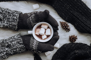 Woman hands in wool gloves holding a cup of hot chocolate with marshmallows decorate with black knitted scarf and pines on monochrome style