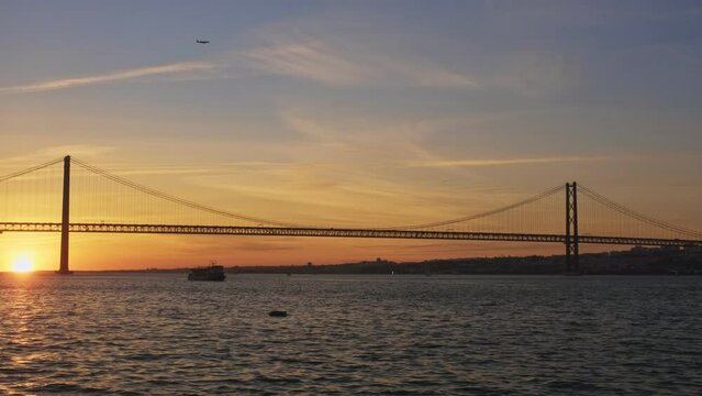 View of 25 de Abril Bridge famous tourist landmark of Lisbon connecting Lisboa and Almada on Setubal Peninsula over Tagus river with boat silhouette on sunset and flying plane. Lisbon, Portugal