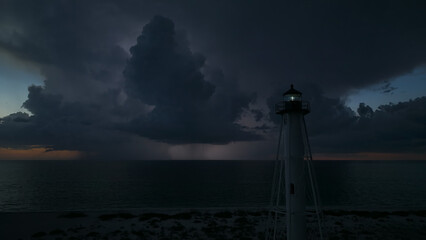 White tall lighthouse on sea shore with blinking light at stormy night for commercial vessels...