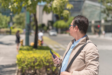A seemingly lost middle aged asian man looks around his surroundings while using a gps map app on his phone. Walking around the city during a hot sunny day.