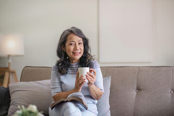 A happy retired 60s Asian woman reading a book while having her coffee on a sofa at home.