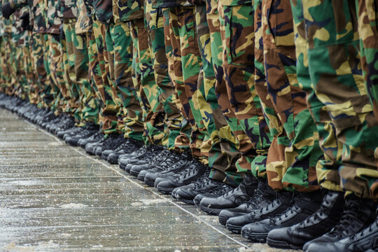 A close-up view of a row of soldiers standing in formation on a wet pavement, wearing black combat boots and camouflage military uniforms