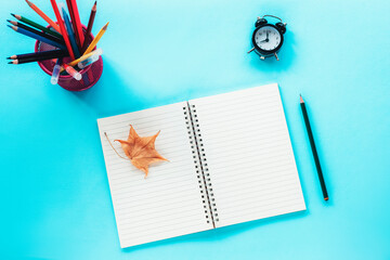 Blank notepad and school supplies, alarm clock, pencil and yellow autumn leaf on blue background. Back to school concept. Top view, flat lay, mock up