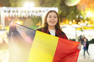 Poster Smiling young traveler girl in warm red and black plaid shirt enjoying walk on traditional street Christmas fair in European town, waving national flag of Belgium .. © JackF