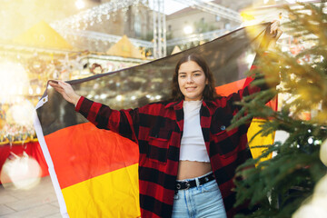 Smiling young traveler girl in white crop top and warm red and black plaid shirt enjoying walk on traditional street Christmas fair in German town, waving national tricolour flag..