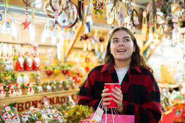 Happy girl walking in colorful Christmas street market, holding paper cup of warm mulled wine
