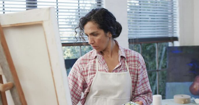 Focused biracial female artist in apron painting on canvas in studio, slow motion