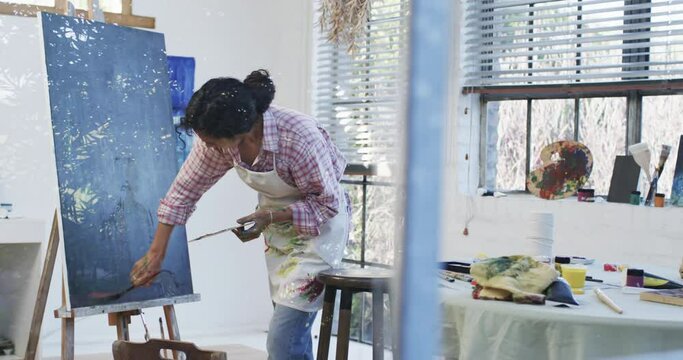 Focused biracial female artist in apron painting on canvas in studio, slow motion