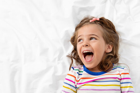 Naklejki Portrait of laughing little girl lying on white bed looking up with funny face