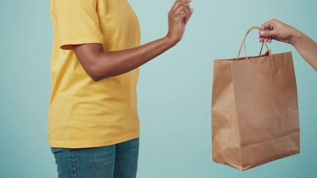 Closeup shot. Delivery woman in yellow uniform giving female client order paper bag with products. Isolated on blue background.