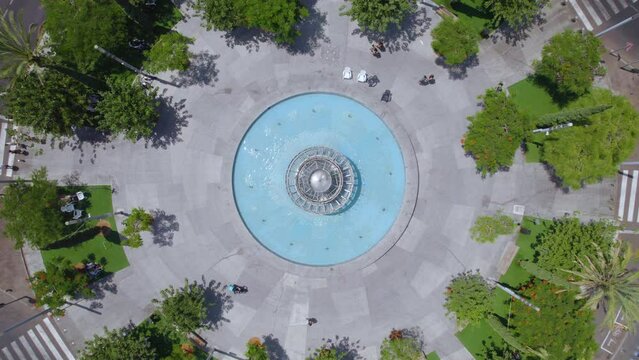 Drone Circling Dizengoff Square, Tel Aviv on a hot summer day as people sit and enjoy the shade of the trees and a cyclist crosses the square