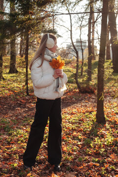 Full lenght of teen cover girl posing with fall leaves in autumn park, pensive looking away. Lovely young lady model in white jacket walking in autumn forest. Leisure activity concept. Copy text space