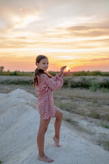 A teenage girl in a summer dress is photographed at sunset.