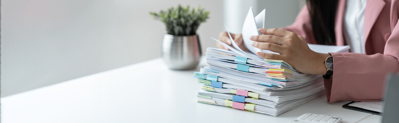 Businesswoman working in stacks of paper files to find and review unfinished documents in document...