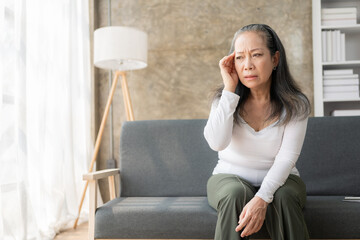 Fototapeta na wymiar Asian senior woman sitting on sofa and having headache in house, hands touching head, lonely asian old woman. Retirement, quarantine and healthcare concept.