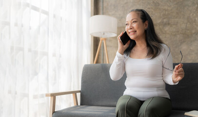 Happy Asian senior woman holding mobile phone for online video call Sitting on the sofa, call family members using mobile applications.