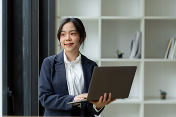 Asian businesswoman standing working with laptop computer, mobile phone, tablet to contact, chat, search internet, news, email, chat while working on finance, marketing, accounting data in office.