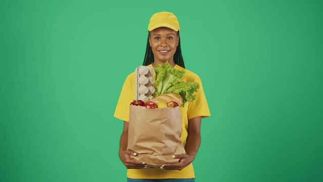 Delivery woman in yellow uniform holding paper bag full with grocery products. Isolated on green background.
