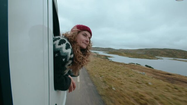 Happy vanlife traveller hang out of car window, catch wind with hand. Concept of travel and adventure. Travel in Scandinavian country. Woman wear traditional wool knit sweater on road trip in Norway