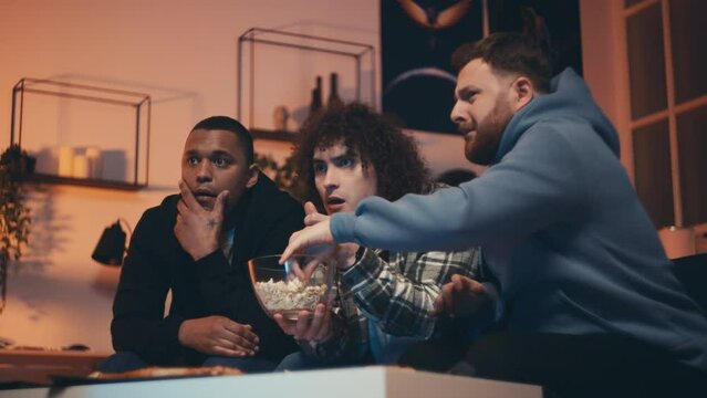 Three male friends watching a scary horror movie at home, eating popcorn