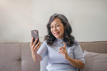 Portrait, Happy 60s retired Asian woman using her phone on a sofa in the living room. chatting,...