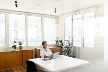 Cheerful African-American female office employee using laptop sitting in the modern coworking space, woman in casual wear and glasses typing indoor, manager on the workplace