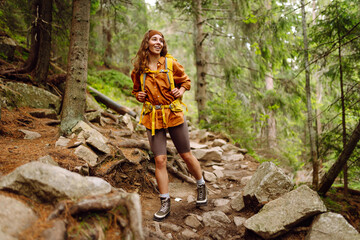 
Traveler with a yellow backpack walks along a hiking path among the mountains in the forest. Curly woman tourist enjoys nature, explores new places.