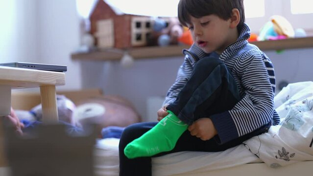 Child putting on sock  seated by bedside. Small boy dressing himself. Kid getting ready to start the day routine