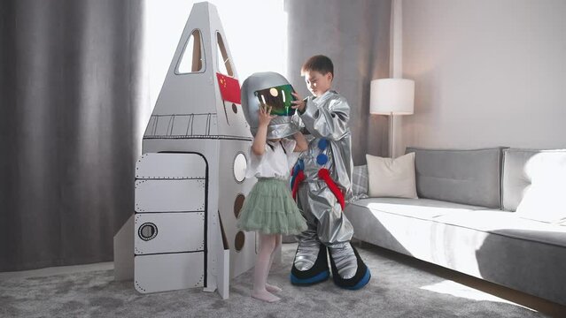 Asian kids play in the living room of the house, a boy in an astronaut costume plays with her sister near the cardboard space shuttle, a cute girl sister puts on a astronaut helmet.