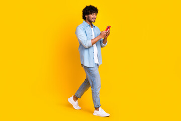 Full length photo of satisfied man wear jeans shirt look at smartphone write post go empty space isolated on yellow color background
