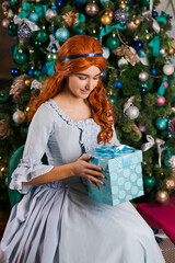 a young woman in a beautiful dress sits near a Christmas tree holding a box with a gift in her hands