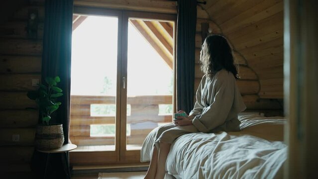 A young woman in a bathrobe enjoying her morning coffee while sitting on the bed.