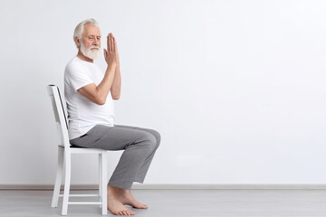 An elderly man practices yoga asana or sports exercises for legs and arms on a chair. The concept of sports in old age, exercise therapy, rehabilitation. Banner. Copy space - obrazy, fototapety, plakaty