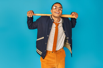 happy and stylish african american student with baseball bat looking at camera on blue background