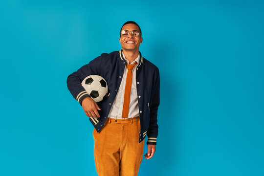 pleased african american student in orange pants, with soccer ball, smiling at camera on blue
