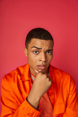 thoughtful and worried african american man in orange shirt touching chest, looking at camera on red