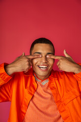 cheerful african american guy obscuring eyes with fingers, orange shirt, red background