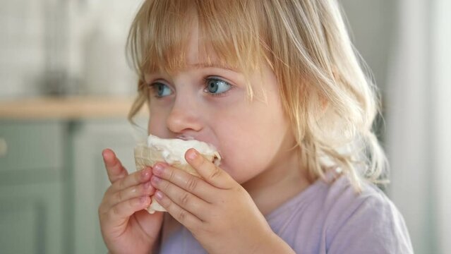 Baby girl enjoying ice cream. Pretty little toddler eating an ice-cream indoors, at home. Dining room background. Small child eats plombir and cream messy on her mouth. Cute kid with tasty sweet food.