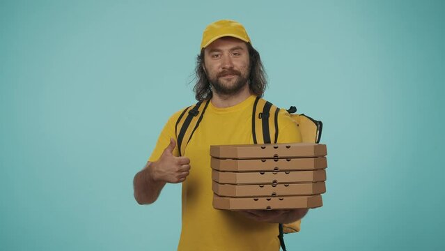 Delivery man in yellow uniform with portable backpack refrigerator holding stack of pizza boxes and shows thumbs up. Isolated on blue background.