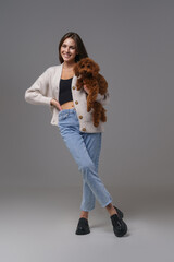 Radiant young brunette in casual wear beaming with happiness while holding her cherished toy poodle, set against a neutral gray studio background