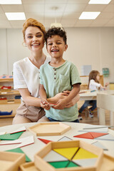 smiling teacher hugging african american kid near didactic materials in montessori school - obrazy, fototapety, plakaty
