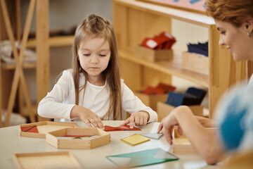 smiling teacher playing with girl and didactic material on table in montessori school - obrazy, fototapety, plakaty