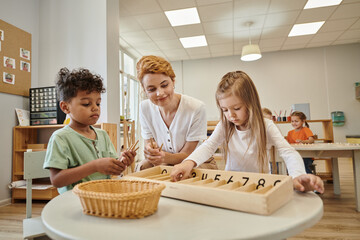 smiling teacher near interracial kids holding wooden sticks during lesson in montessori school