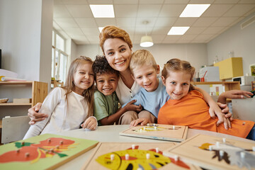 smiling teacher hugging multiethnic kids near didactic materials on table in montessori school
