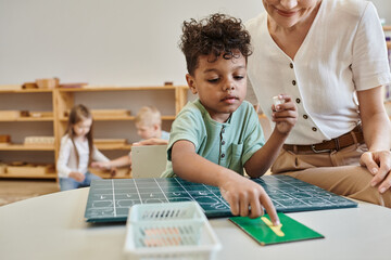 cheerful teacher observing african boy pointing at number near chalkboard, learning through play